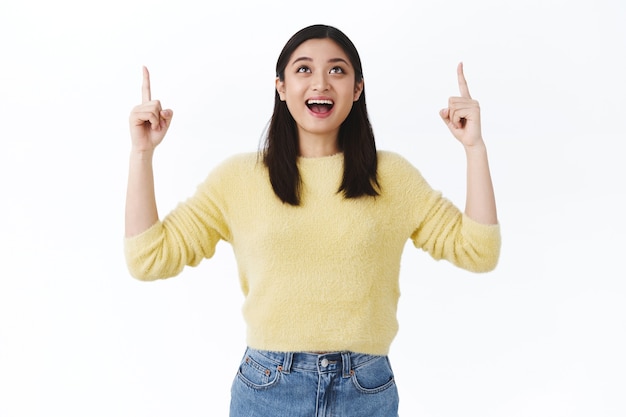 Dreamy lucky asian girl feeling excited and happy, receiving great chance, pointing finger up at dream that came true, smiling upbeat standing white wall astonished, rejoicing good news