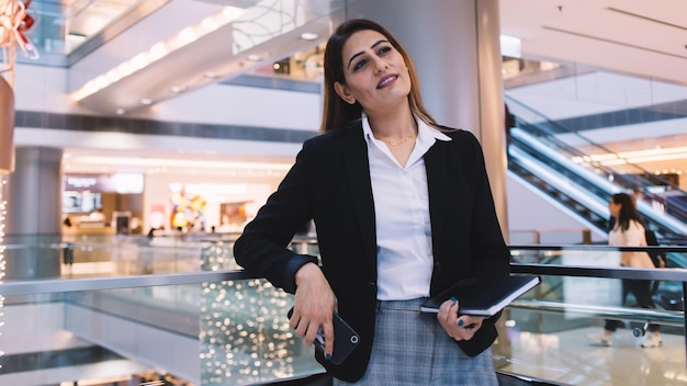 Dreamy manager in strict suit standing in bright shopping mall and holding book and smartphone