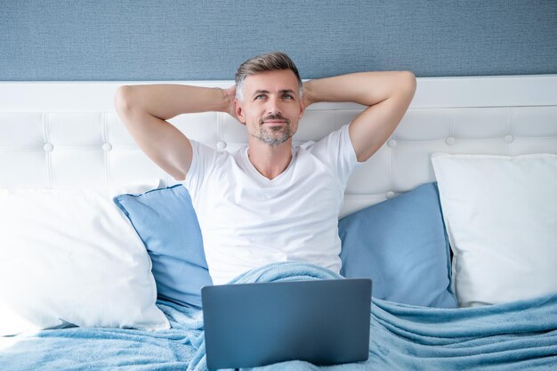 Dreamy mature man relax in bed with laptop