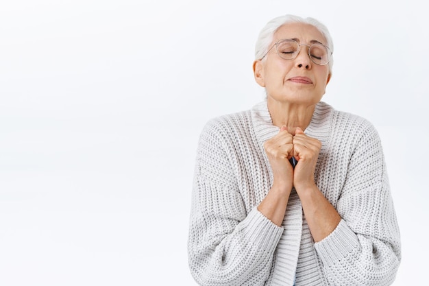 Dreamy and overwhelmed elegant senior woman recall youth memories, hold hands together tight near chest