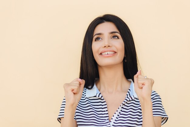 Sognante donna positiva con i capelli lisci neri, tiene le mani nei pugni