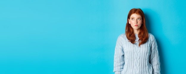 Dreamy redhead girl thinking or making decision looking at upper left corner logo standing against b