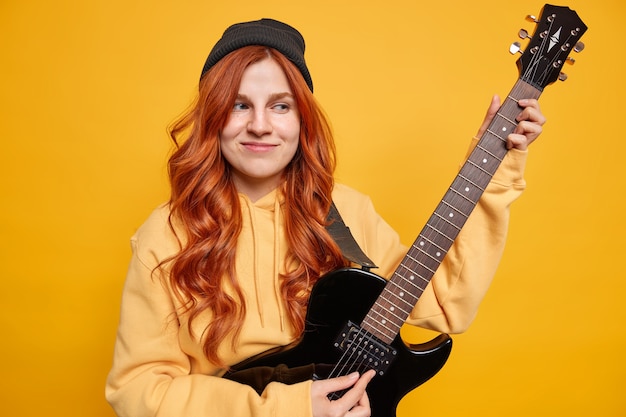 Dreamy satisfied female teenager musician plays electric guitar wants to become professional guitarist wears hoodie and hat has long red hair 