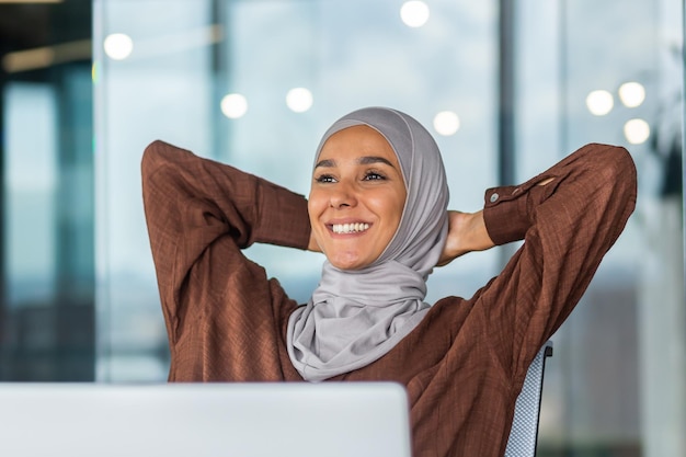 A dreamy and smiling young arab woman in a hijab is sitting in the office on a chair hands behind