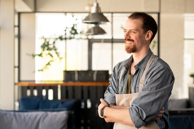 Dreamy successful young bearded coffee shop owner in apron standing with crossed arms and looking into distance