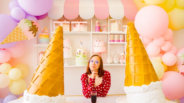 Мечтательная женщина пьет освежающий напиток возле больших декоративных конусов мороженого