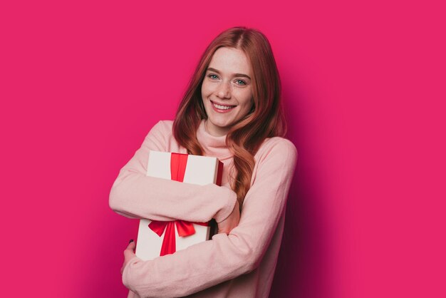 Dreamy woman hugging her surprise gift and smiling while looking at camera standing on pink backgrou...