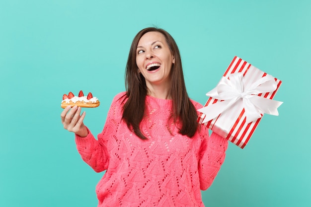 Dreamy woman in pink sweater looking up, hold eclair cake, red striped present box with gift ribbon isolated on blue background. Valentine's, Women's Day, birthday holiday concept. Mock up copy space.