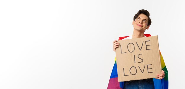 Dreamy young queer person smiling and looking at upper left corner holding love is love sign for pride parade wearing Rainbow flag white background