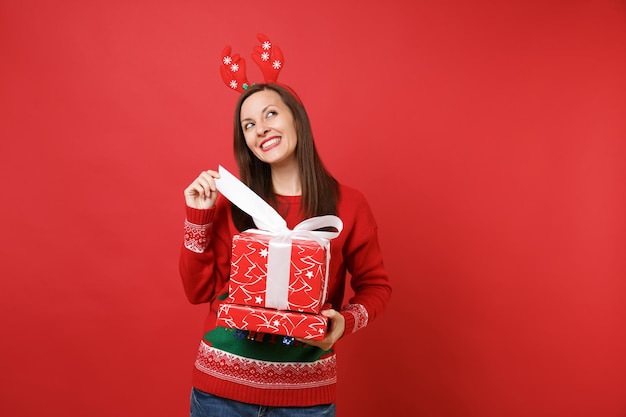 Dreamy young Santa girl in fun decorative deer horns on head looking up, opening red boxes with gifts presents isolated on bright red background. Happy New Year 2019 celebration holiday party concept.