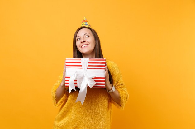 Dreamy young woman in birthday hat looking up, holding red box with gift, present enjoying holiday isolated on bright yellow background. People sincere emotions, lifestyle concept. Advertising area.