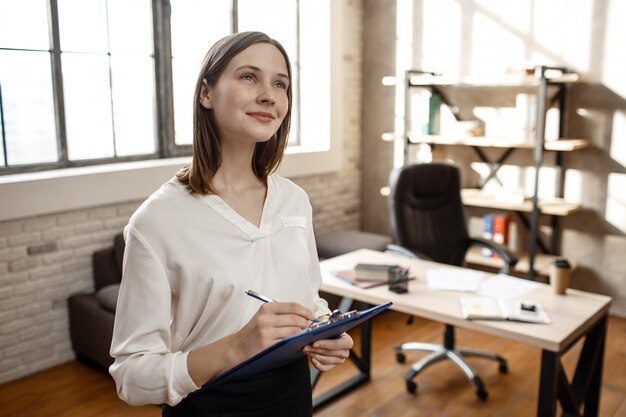 Dreamy young woman stand in room aloone and look up. She write and smile. Empty room. Daylight.