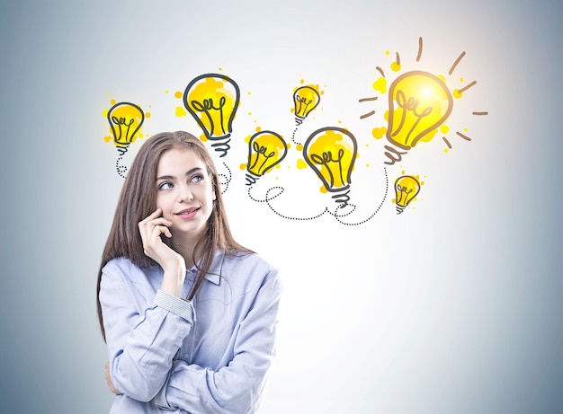 Photo dreamy young woman with brown hair wearing a blue shirt and business suit pants is looking upwards and thinking. yellow light bulbs on a gray wall. an idea concept