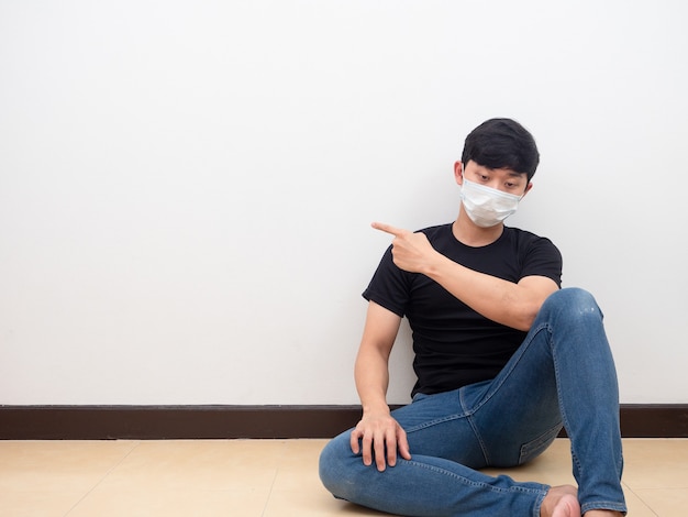 Drepressed Asian man wearing mask sit on the floor point finger at space white background