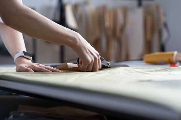 洋裁の手は、はさみでカットされた生地素材を使用して、仕立てのワークショップで作業プロセスのクローズアップを縫います