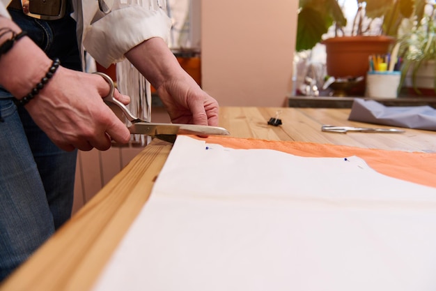 양장점은 재단사 작업장에서 주황색 새틴 천으로 재봉 패턴을 재단 가위를 사용하여 새 의류 클로우즈업 패션 디자이너의 손을 시작합니다.