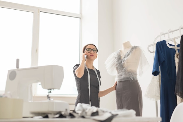 Dressmaker, tailor, fashion and showroom concept - beautiful young designer is talking on the mobile phone and smiling while examining her work in dressmaking studio.