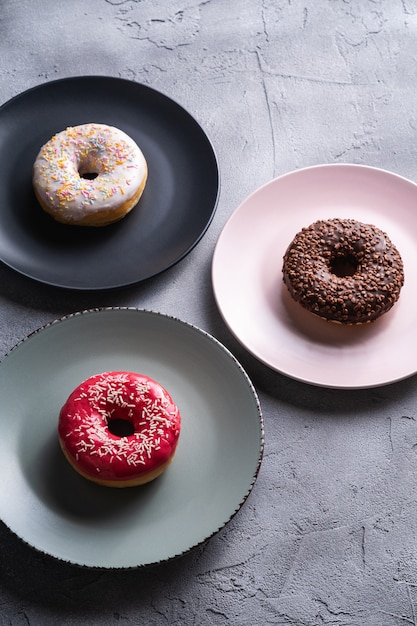 Drie donuts op borden, chocolade, roze en vanille donut met hagelslag, zoet geglazuurd dessertvoedsel op betonnen structuur, hoekmening