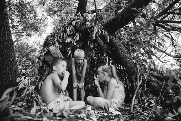 Drie dorpskinderen spelen in een hut die ze zelf hebben gebouwd van bladeren en takjes Houten huis in het bos Zwart-wit fotografie