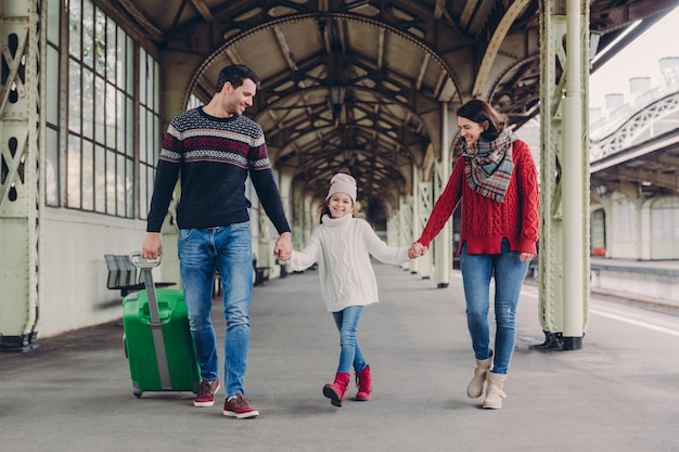 Drie familieleden op treinstation gelukkige moeder, dochter en vader hebben positieve gezichtsuitdrukkingen wacht op trein op perron genieten van saamhorigheid blije passagiers hebben reis naar het buitenland