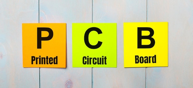 Drie gekleurde stickers met de tekst PCB Printed Circuit Board een lichtblauwe houten ondergrond