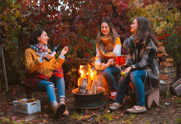 Drie gelukkige, vrolijke beste vrienden die buiten in de herfsttuin zitten bij het kampvuur met een fles rode wijn