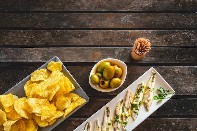 Drie gerechten met chips, geplukte ansjovis en groene olijven op een donkere houten tafel met tandenstokers. Typische Spaanse snacks.