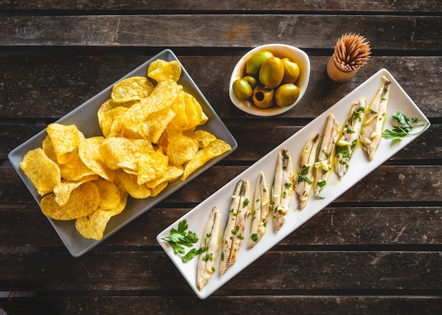 Drie gerechten met chips, geplukte ansjovis en groene olijven op een donkere houten tafel met tandenstokers. Typische Spaanse snacks.