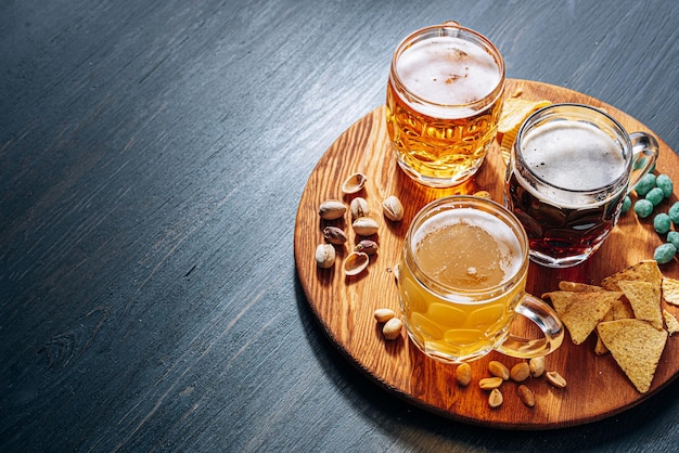 Drie glazen duur ambachtelijk bier, klassiek en ongefilterd en donker in een glas op tafel met een snack van pinda en pistache chips en nacho's