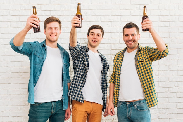 Foto drie glimlachende mannelijke vriend die bierfles opheffen die zich tegen witte bakstenen muur bevinden