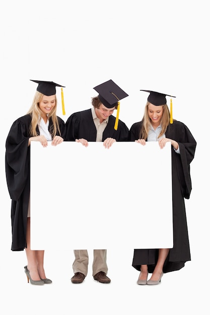 Foto drie glimlachende studenten in gediplomeerde robe die een leeg teken houdt