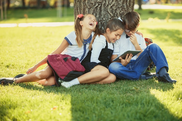 Drie glimlachende tienerschoolvrienden die in park op gras zitten en samen nieuw tabletspel spelen. Verschillende emoties op hun gezichten