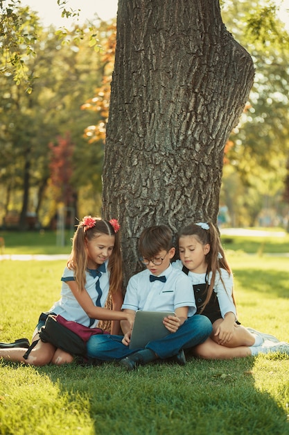 Drie glimlachende tienerschoolvrienden die in park op gras zitten en samen nieuw tabletspel spelen. verschillende emoties op hun gezichten