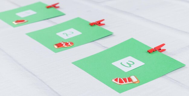Drie groene vellen papier met nummers 1 2 3 en kerstdecor bevestigd met rode mini wasknijpers op een witte bakstenen muur