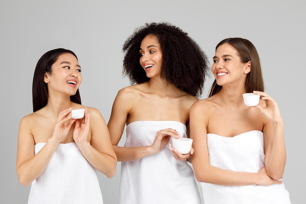 Drie jonge diverse vrouwen die cosmetische natuurlijke gezichtscrèmepotten vasthouden en voor hun huid zorgen