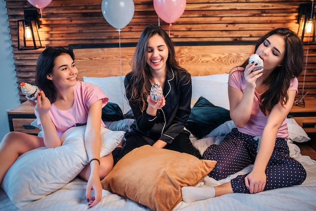 Drie jonge gelukkige vrouwen zitten op bed. Twee van hen kijken naar akes en glimlachen. De derde eet het op. Taart is heerlijk. Meisjes zijn in de kamer. Sommige ballonnen lopen achter.