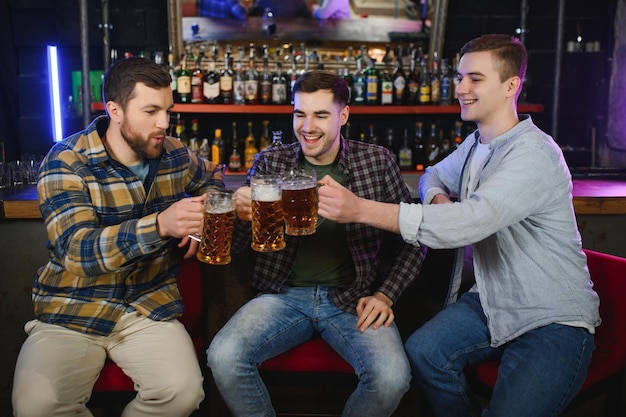 Drie jonge mannen in vrijetijdskleding glimlachen en kletteren samen glazen bier terwijl ze aan de toog in de pub zitten