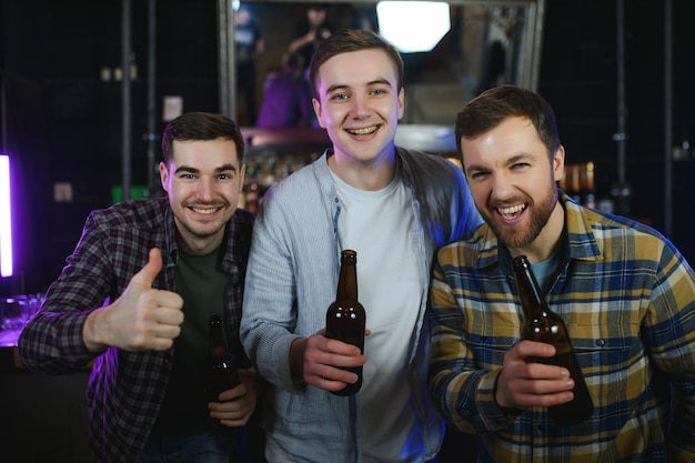 Drie jonge mannen in vrijetijdskleding glimlachen terwijl ze flessen bier vasthouden terwijl ze in de buurt van de toog in de pub staan