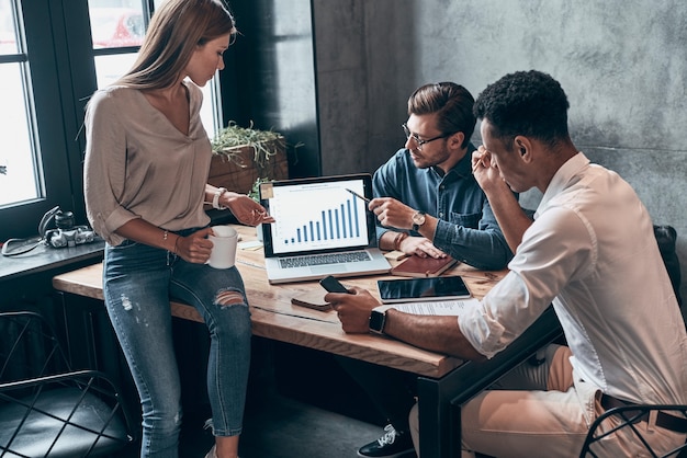 Drie jonge mensen in slimme vrijetijdskleding die gegevens op laptop analyseren terwijl ze op kantoor werken