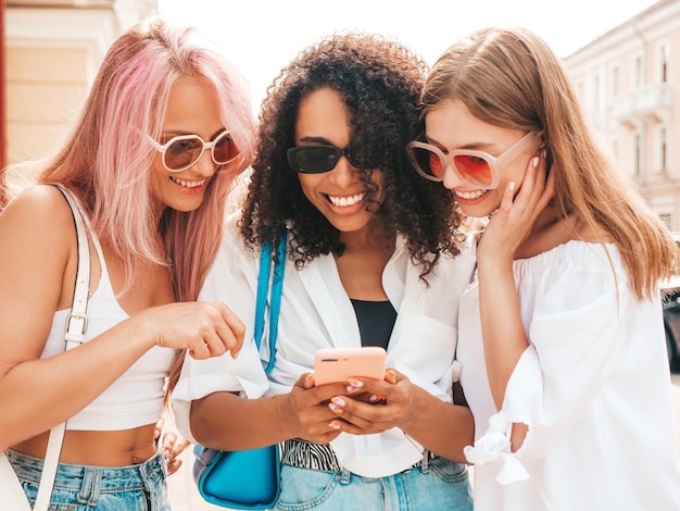 Drie jonge mooie glimlachende hipster vrouw in trendy zomerkleren Sexy zorgeloze multiraciale vrouwen poseren in de straat Positieve modellen in zonnebril kijken naar smartphone scherm Telefoon apps gebruiken