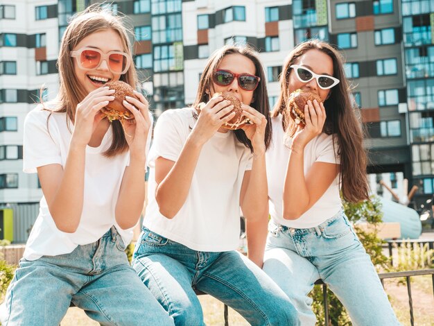 Drie jonge mooie lachende hipster vrouw in trendy zomer dezelfde kleding Sexy zorgeloze vrouwen poseren in de straat Positieve modellen plezier in zonnebril houden van sappige hamburger en hamburger eten