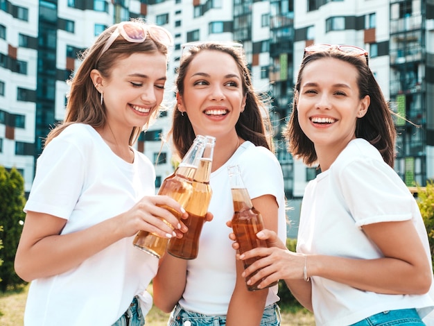 Drie jonge mooie lachende hipster vrouw in trendy zomer dezelfde kleren Sexy zorgeloze vrouwen poseren in de straat Positieve modellen plezier in zonnebril Drinkfles bier Oktoberfest