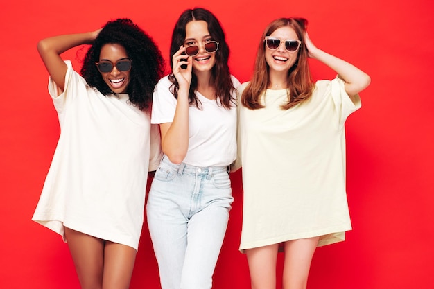 Drie jonge mooie lachende internationale hipster vrouw in trendy zomerkleren sexy zorgeloze vrouwen poseren in de buurt van rode muur in studio positieve modellen plezier in zonnebril Concept van vriendschap