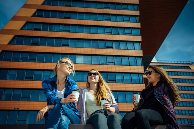 Drie jonge mooie vrouwen zitten op een bankje in een straat in de stad, drinken koffie uit een wegwerpbril, praten en lachen