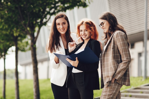 Drie jonge stijlvolle vrouwen die buitenshuis werken en glimlachen, houden een map met documenten vast