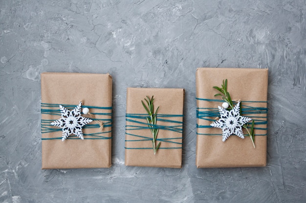 Drie kerstcadeaus in ambachtelijk papier gewikkeld in blauwe draad, met witte sneeuwvlokken en rozemarijntakken op een grijze betonnen achtergrond.