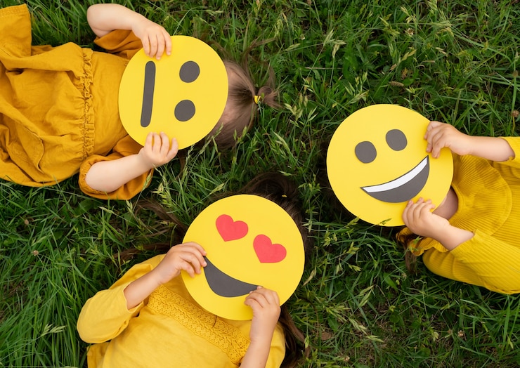 Foto drie kinderen die op het gras liggen, houden kartonnen emoticons vast met verschillende emoties