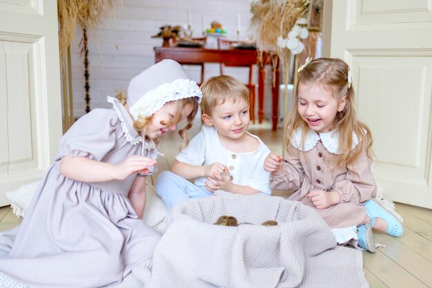 drie kinderen kleine blonde meisjes in vintage jurk en schattig kleine jongen in de woonkamer landhuis op de lente dag spelen met baby eenden eendjes gelukkige jeugd eenvoudige gezellige leven gelukkige paas