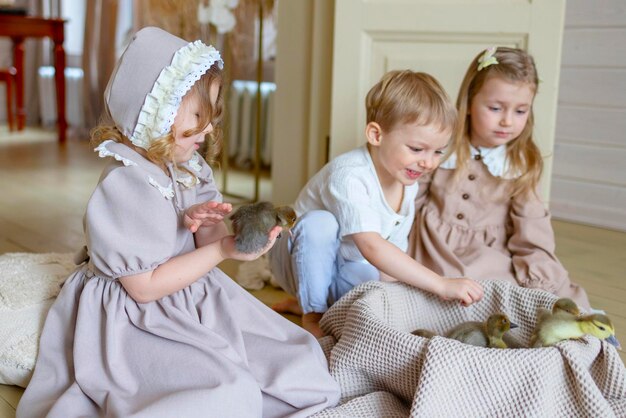 drie kinderen kleine blonde meisjes in vintage jurk en schattig kleine jongen in de woonkamer landhuis op de lente dag spelen met baby eenden eendjes gelukkige jeugd eenvoudige gezellige leven gelukkige paas