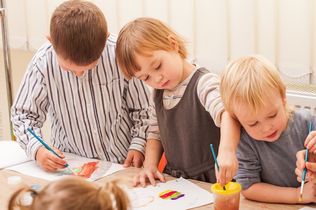 Drie kinderen leren schilderen met een penseel en aquarellen op papier in de kleuterschool